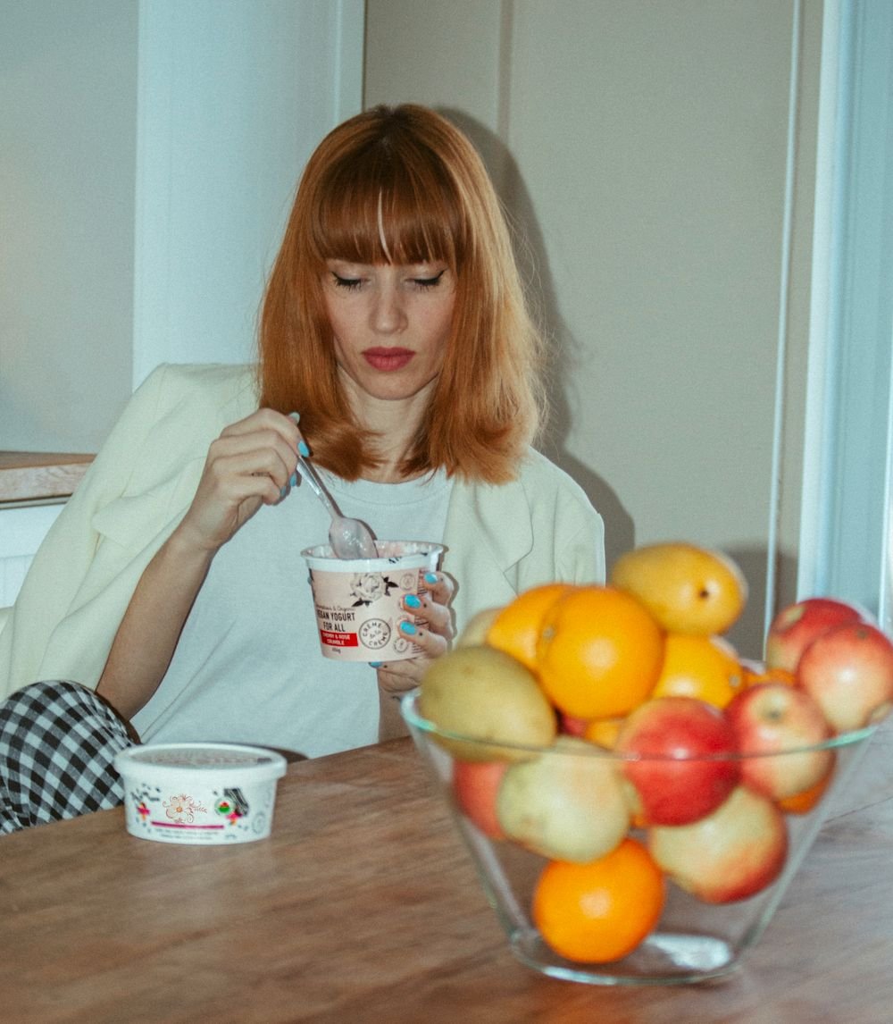 Mulher à mesa saboreando iogurte de colher, ao lado de frutas