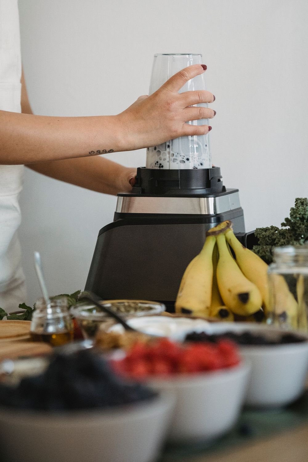 Mãos de uma mulher preparando uma bebida no liquidificador, com frutas sobre a bancada